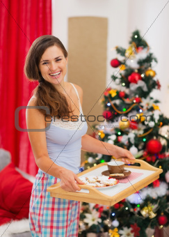 Happy woman in pajamas holding bed table with snacks in front of Christmas tree