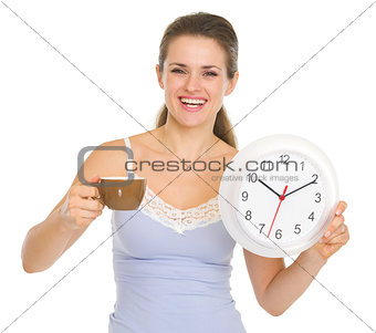 Happy young woman in pajamas with cup of coffee and clock