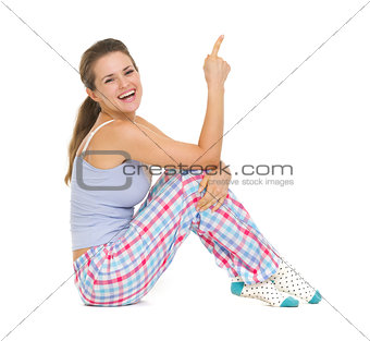 Smiling young woman in pajamas sitting on floor and pointing on 