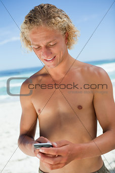 Smiling blonde man standing on the beach while sending a text 