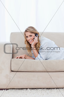 A smiling woman talking on her mobile phone is lying on a couch