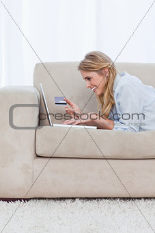 A woman resting on her elbows is holding a bank card and typing 