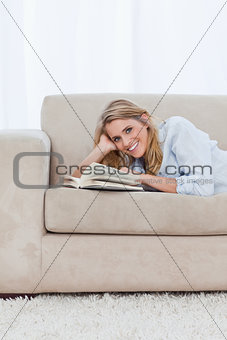 A woman looking at the camera is lying on a couch with a book