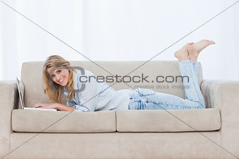 A smiling woman lying on a couch using a laptop