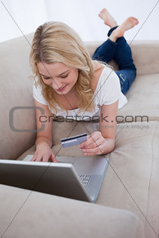 A woman lying on a couch is holding her bank card and typing on 