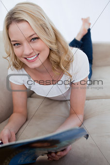 A smiling woman is lying on a couch and holding a magazine