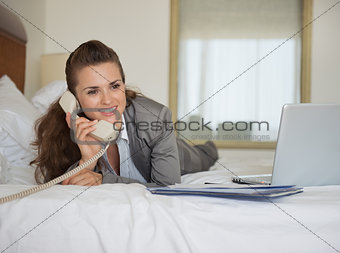 Business woman laying in bed with laptop and talking phone in ho
