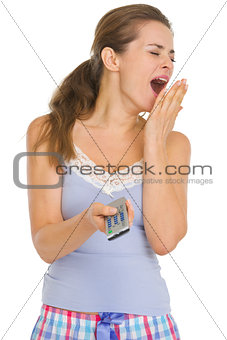 Young woman in pajamas with TV remote control yawing