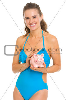 Smiling young woman in swimsuit holding piggy bank