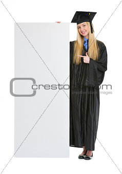 Happy young woman in graduation gown pointing on blank billboard