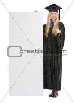 Happy young woman in graduation gown showing blank billboard and