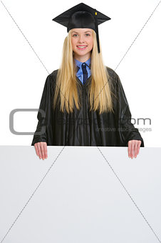 Smiling young woman in graduation gown showing blank billboard