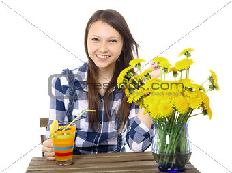 Girl teenager, caucasian appearance, brunette, wearing a plaid shirt, holding a glass of drink. On the table is a blue vase,  with a bouquet of yellow wildflowers, dandelions.