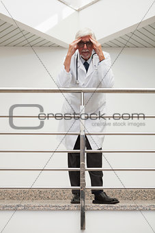 Stressed doctor leaning on  railing with hands on forehead in hospital corridor