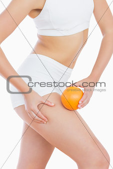 Fit woman squeezing fat on thigh as she holds orange