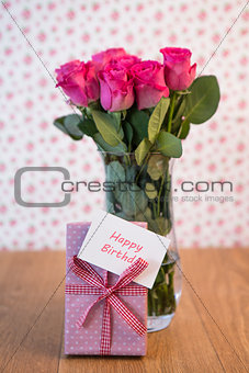 Bunch of pink roses in vase with pink gift leaning against it and happy birthday card