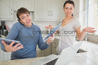 Busy man using two tablets and laptop with wife holding hands up questioning
