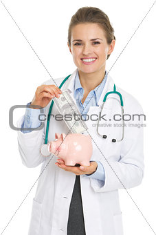 Smiling doctor woman putting dollars banknote in piggy bank
