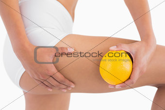 Slim woman squeezing cellulite skin on thigh as she holds orange