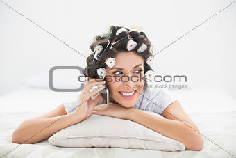Happy brunette in hair rollers lying on her bed making a phone call