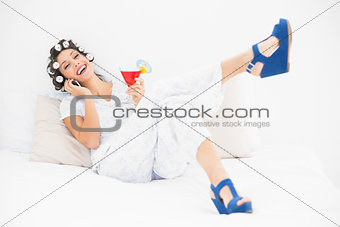 Brunette in hair rollers and wedge shoes having a cocktail making a call on bed