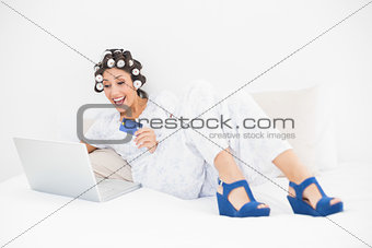 Excited brunette in hair rollers and wedge shoes using her laptop for shopping online