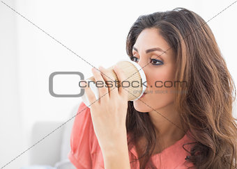Smiling brunette sitting on her sofa sipping from disposable cup