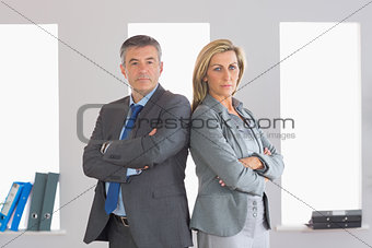 Two unsmiling businesspeople looking at camera standing back to back with crossed arms