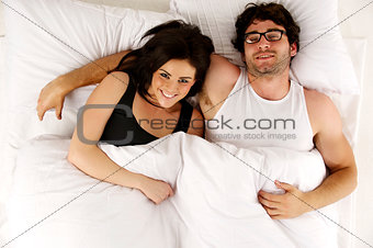 Man and woman laid in white bed looking up at the camera smiling