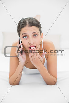 Astonished young brown haired model in white pajamas making a phone call