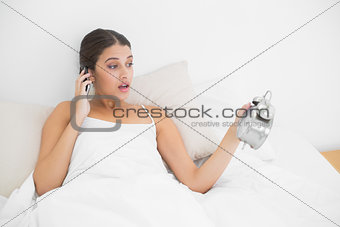 Surprised young brown haired model in white pajamas on the phone looking at her alarm clock