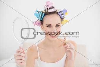 Charming natural brown haired woman in hair curlers holding a mirror and an eyelash curler