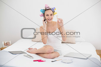 Cute natural brown haired woman in hair curlers making a phone call and holding an eyelash curler