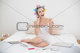 Surprised natural brown haired woman in hair curlers applying gloss while being on the phone