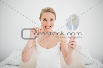 Natural happy blonde holding mirror and using eyelash curler