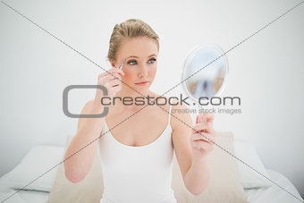 Natural serious blonde holding mirror and using tweezers