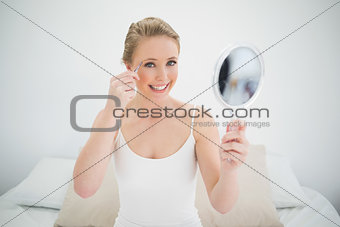 Natural smiling blonde holding mirror and using tweezers