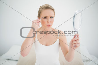 Natural frowning blonde holding mirror and using tweezers
