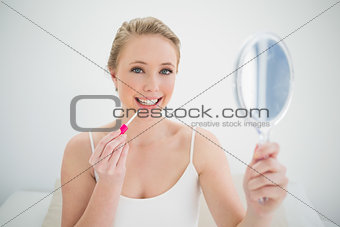 Natural smiling blonde holding mirror and applying lip gloss
