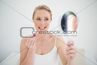 Natural happy blonde holding mirror and applying lip gloss