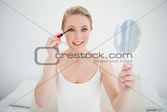 Natural smiling blonde holding mirror and applying mascara