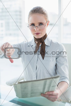 Blonde serious businesswoman holding newspaper and showing thumb down