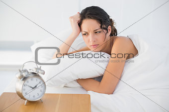 Frowning casual brunette in white pajamas looking at her alarm clock