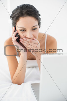 Happy casual brunette in white pajamas making a phone call and looking at her laptop