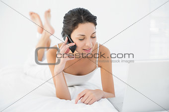 Calm casual brunette in white pajamas making a phone call and looking at her laptop
