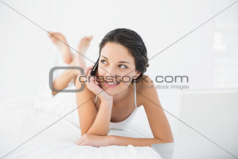 Smiling casual brunette in white pajamas making a phone call and looking away