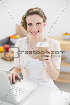 Gorgeous woman using her notebook in her kitchen holding a disposable cup