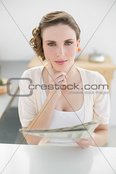 Gorgeous thinking woman sitting in her kitchen holding newspaper