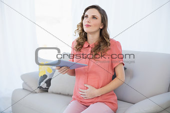 Thinking pregnant woman holding her tablet while sitting on a couch in the living room