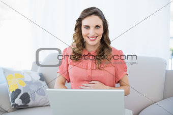 Cheerful pregnant woman using her notebook while sitting on couch